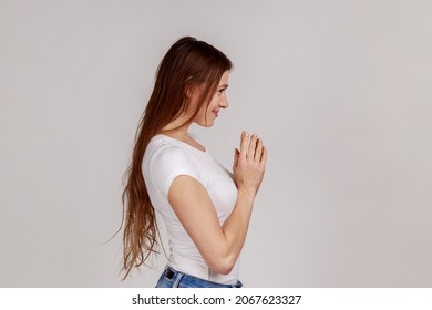 Side view portrait of scheming devious woman clasping hands and thinking over tricky plan, having sly cunning idea to prank, wearing white T-shirt. Indoor studio shot isolated on gray background.