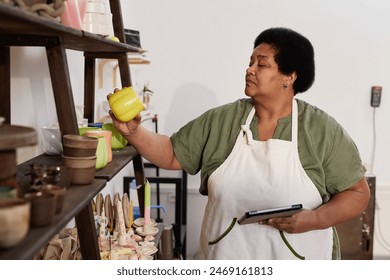 Side view portrait of senior African American woman inspecting glazed pottery pieces on shelf in art studio - Powered by Shutterstock