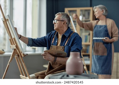 Side view portrait of senior man as artist painting on easel in art studio enjoying creative hobby copy space - Powered by Shutterstock