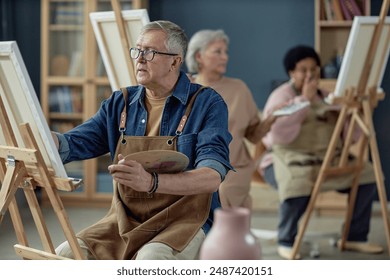 Side view portrait of senior man painting on easel in art studio with group of elderly people in background copy space - Powered by Shutterstock