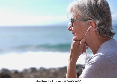 Side view portrait of senior smiling woman sitting on the beach listening to music with earphones. Gray haired woman relaxing looking at horizon over sea.