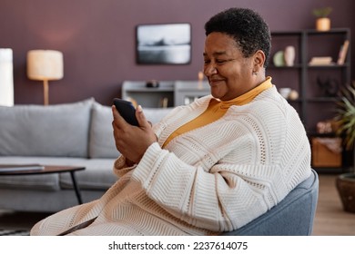 Side view portrait of senior woman using smartphone while relaxing in chair in cozy home setting and smiling cheerfully - Powered by Shutterstock