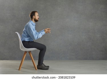 Side view portrait of a serious businessman sitting tense at an imaginary desk with a computer. Man keeps his hands outstretched as if typing on a keyboard and working on a laptop. Gray background.