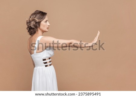 Side view portrait of serious middle aged woman with wavy hair showing stop palm gesture, prohibition sign, wearing white dress. Indoor studio shot isolated on light brown background.