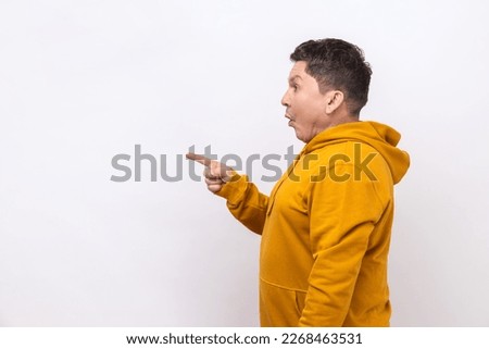 Side view portrait of shocked man pointing finger away at empty space for advertisement, looking aside with open mouth, wearing urban style hoodie. Indoor studio shot isolated on white background.