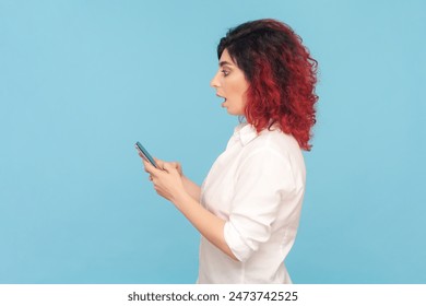 Side view portrait of shocked surprised woman with fancy red hair holding smart phone with big eyes, checking social networks, wearing white shirt. Indoor studio shot isolated on blue background. - Powered by Shutterstock