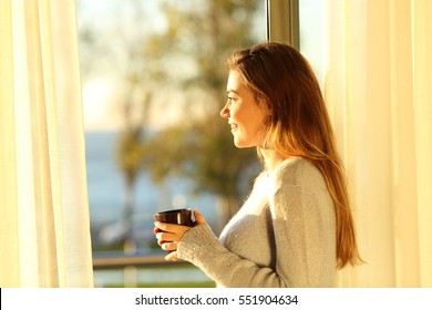 Side view portrait of a single relaxed girl looking the sea outside through a window and holding a coffee mug at sunset in the living room at home