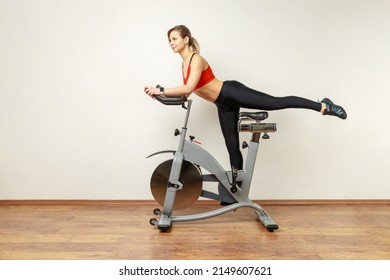 Side View Portrait Of Slim Attractive Woman Standing On Exercise Bike With Raised Leg, Cardio Workout, Wearing Sports Tights And Red Top. Indoor Studio Shot On Gray Wall Background.