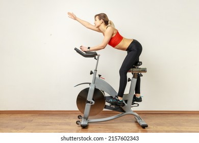 Side view portrait of slim woman riding exercise bike and raised arm, doing sport exercises during cardio workout, wearing sports tights and red top. Indoor studio shot on gray wall background.