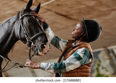 Side view portrait of smiling African American woman stroking black horse in indoor riding arena at practice - Powered by Shutterstock