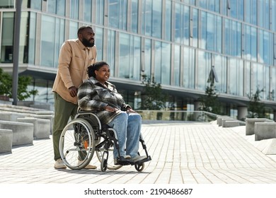 Side view portrait of smiling black man assisting partner in wheelchair outdoors in city against glass building architecture, copy space