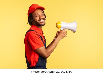 Side View Portrait Of Smiling Happy Handyman Wearing Blue Overalls And Red Cap, Holding Megaphone, Looking At Camera With Toothy Smile. Indoor Studio Shot Isolated On Yellow Background.