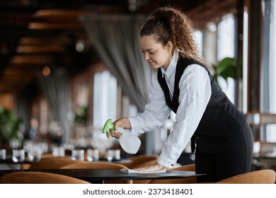 Side view portrait of smiling young woman as server wearing classic uniform and cleaning dining tables in restaurant, copy space - Powered by Shutterstock