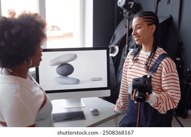 Side view portrait of smiling young woman as female photographer holding camera and talking to friend or colleague during product photography photoshoot in studio - Powered by Shutterstock