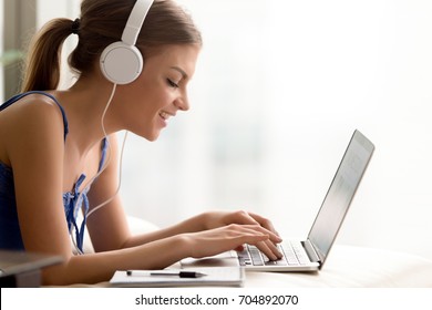 Side View Portrait Of Smiling Young Woman In Headphones Typing On Laptop While Taking Online Course Or Personal Training For Self-education, Qualification Increase. Happy Lady Studying In Internet 