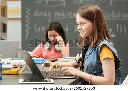 Side view portrait of smiling young girl with prosthetic arm using laptop during IT class for children in school copy space