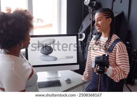 Side view portrait of smiling young woman as female photographer holding camera and talking to friend or colleague during product photography photoshoot in studio
