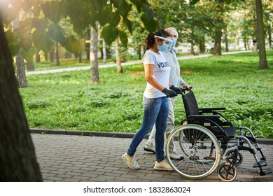 Side View Portrait Of Social Worker And Older Man Walking In The City Park Or Wood With Wheelchair