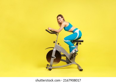 Side View Portrait Of Sporty Attractive Woman Training At Home, Using Excise Bike, Looking Smiling At Camera, Wearing Blue Sportswear. Indoor Studio Shot Isolated On Yellow Background.