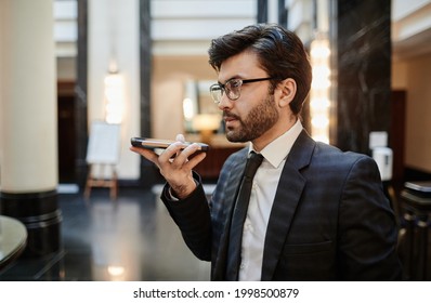 Side View Portrait Of Successful Businessman Recording Voice Message To Smartphone While Standing In Hotel Lobby, Copy Space