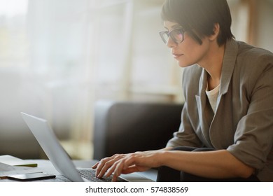 Side View Portrait Of Successful Female Executive Wearing Short Haircut And Glasses Leaning In To Desk While Typing At Laptop Working In Modern Sunlit Office