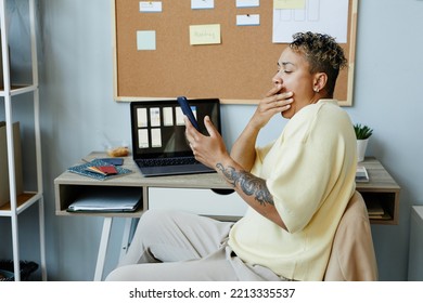 Side View Portrait Of Tattooed Black Woman Yawning At Office Workplace And Scrolling Social Media Via Phone