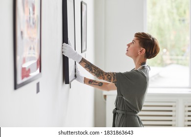 Side view portrait of tattooed creative woman hanging paintings on wall while planning art gallery exhibition, copy space