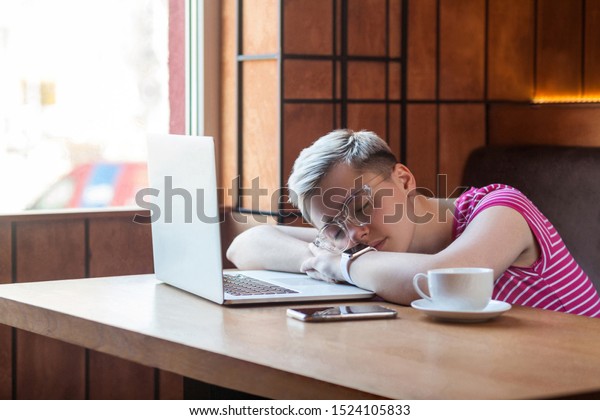 Side view portrait of tired kind beautiful\
young bussineswoman with blonde short hair in pink t-shirt and\
eyeglasses is sitting in cafe and fall asleep on the table. Indoor,\
healthy lifestyle
