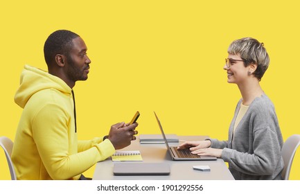 Side View Portrait Of Two Contemporary Business People Sitting Opposite Each Other At Desk During Meting And Smiling Against Yellow Background