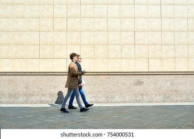 Side View Portrait Of Two Modern Handsome Men Wearing Autumn Coats Walking In City Street Against Beige Concrete Wall