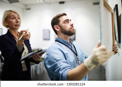 Side view portrait of two museum workers hanging pictures in art gallery, copy space - Shutterstock ID 1303281124