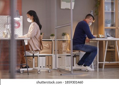 Side View Portrait Of Two People Wearing Masks In Office While Working At Desks In Separate Cubicles Post Pandemic, Copy Space