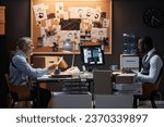 Side view portrait of two police detectives sitting at desk in office on opposite sides studying case investigation