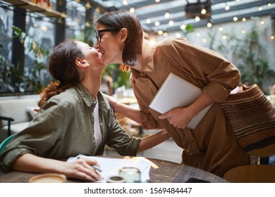 Side view portrait of two young women kissing each other on cheek during meeting in cafe on outdoor terrace decorated with lights