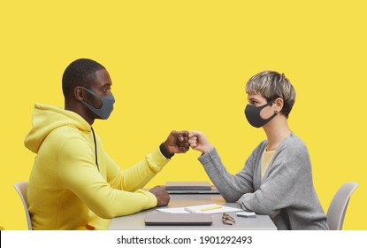 Side view portrait of two young business people wearing masks and bumping fists while sitting opposite each other at desk during meting against yellow background