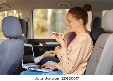 Side View Portrait Of Woman Working On Laptop And Using Voice Assistant For Searchng Information While Sitting With Her Baby Daughter In Safety Chair On Backseat Of The Car.