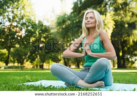 Side view portrait of a yoga on fitness mat in green public park forest. Mature beautiful woman practicing meditation relaxing feeling zen-like