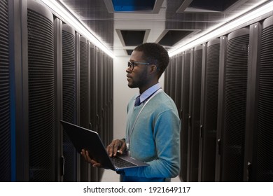 Side view portrait of young African American data engineer working with supercomputer in server room and holding laptop, copy space