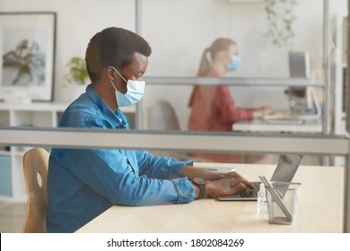 Side View Portrait Of Young African-American Man Wearing Mask And Using Laptop While Sitting At Desk In Cubicle At Post Pandemic Office, Copy Space