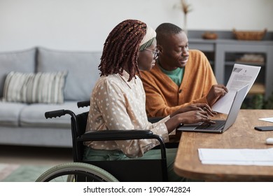 Side view portrait of young African-American woman using wheelchair working from home with husband helping her copy space