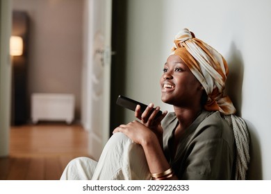 Side view portrait of young African-American woman recording voice message while sitting on floor at home, copy space