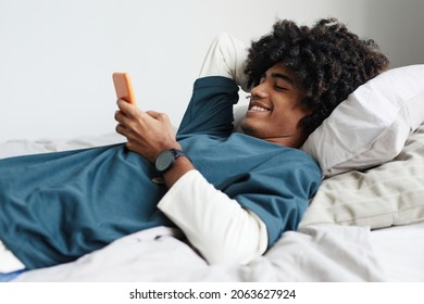 Side view portrait of young African-American man lying on bed at home and smiling while using smartphone
