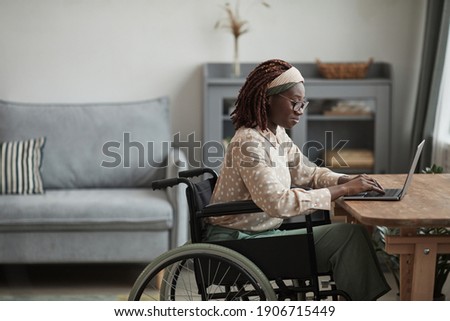 Side view portrait of young African-American woman using wheelchair while working from home in minimal grey interior, copy space