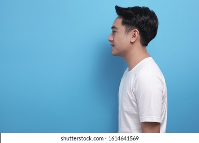 Side View Portrait Of Young Asian Man, Wearing White Shirt Against Blue Background