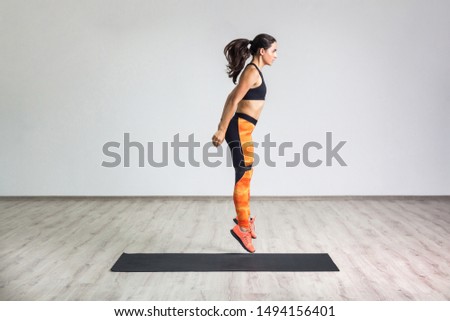 Side view portrait of young athletic healthy woman wearing black top and orange leggings jumping with elastic resistance band, air fly. Isolated, white wall, indoor, workout concept, looking away