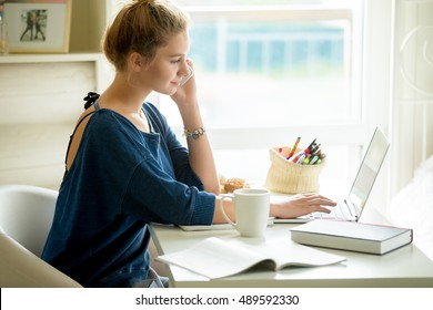 Side view portrait of young beautiful casual woman making call, using smartphone, talking on phone while sitting at modern workplace with laptop, books, coffee and cookies in small home office