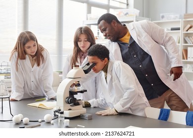 Side View Portrait Of Young Black Girl Looking Into Microscope With While Doing Experiments With Group Of Children In School Chemistry Lab, Copy Space