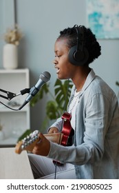 Side View Portrait Of Young Black Woman Singing To Microphone And Playing Guitar In Home Recording Studio