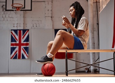 Side view portrait of young Black woman as female basketball player preparing for match sitting on bench in indoor court with flags in background copy space - Powered by Shutterstock