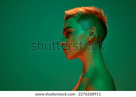 Side view portrait of young blonde girl with short hair and bare shoulders posing against green studio background in orange neon light. Cyberpunk culture. Concept of technology, modern fashion